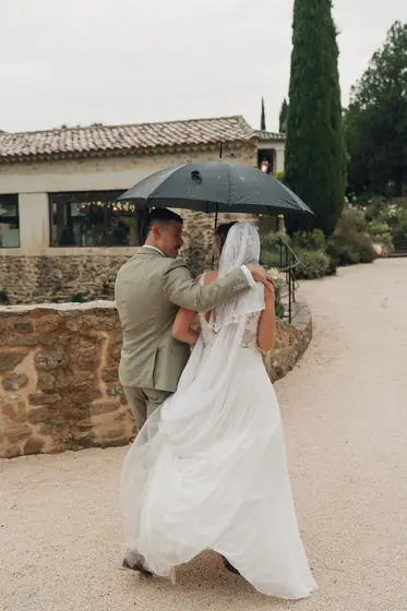 Les mariés marchent sous un parapluie noir sur un chemin de gravier devant une bastide