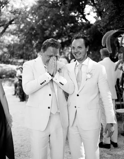 Deux mariés en costume blanc, l'un se cache le visage, ému