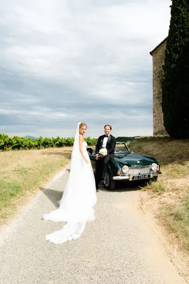 Mariée au long voile devant une voiture ancienne verte, sur un chemin de vignes